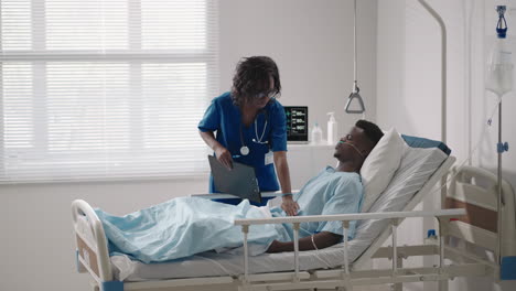 A-black-female-doctor-is-talking-to-a-black-male-patient-lying-on-a-hospital-bed-and-connected-to-an-acid-supply